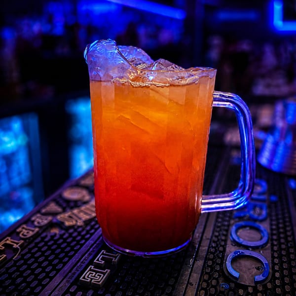 Vibrant orange and red cocktail with ice in a clear pitcher on a bar mat, illuminated by blue neon.