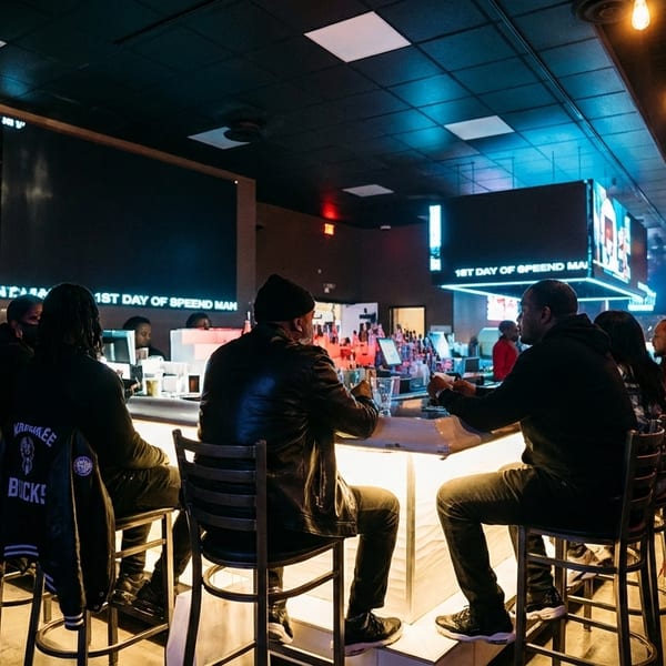 Patrons seated at a dimly lit bar with glowing screens and warm pendant lights overhead.