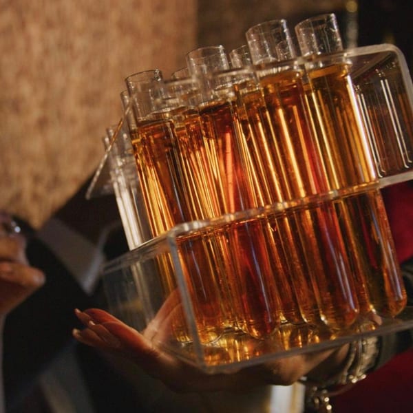 Hand holding a clear tray of amber liquid shots in test tubes.