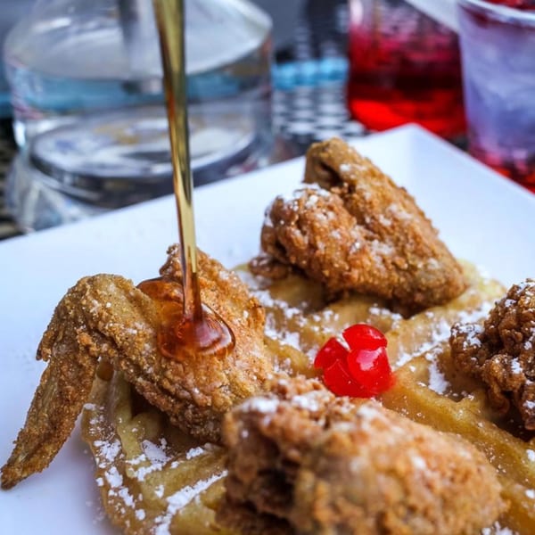 Syrup pouring over fried chicken and waffles with powdered sugar and a cherry on a white plate.