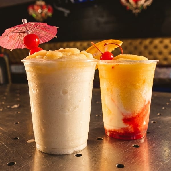 Two colorful frozen drinks with fruit garnishes and a pink umbrella on a metal table.