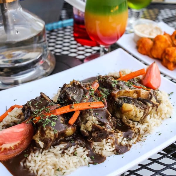 Oxtail stew with rice, carrots, and tomato on a white plate, with drinks and appetizers.